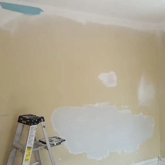 Patchwork spots on yellow wall that is being prepped to paint. Sample of blue paint in upper corner and ladder up against the wall.