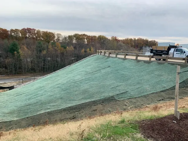 Workers covering a large dirt slope with green erosion control matting beside a wooded area.