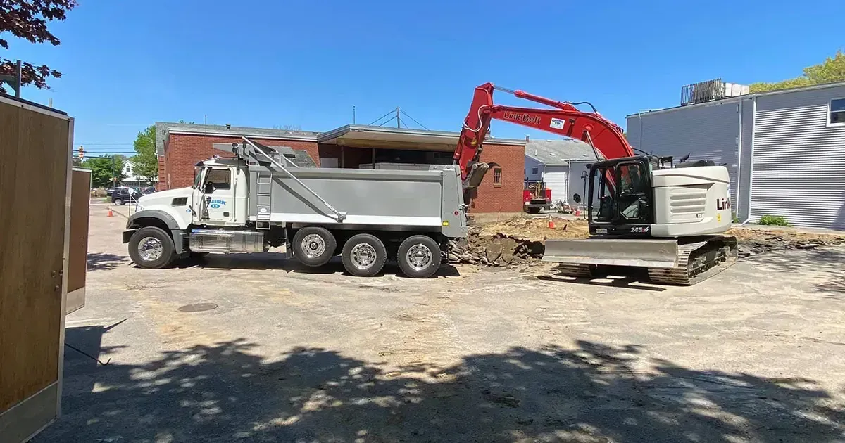 An excavator loading a dump truck with debris from a building demolition under a blue sky.