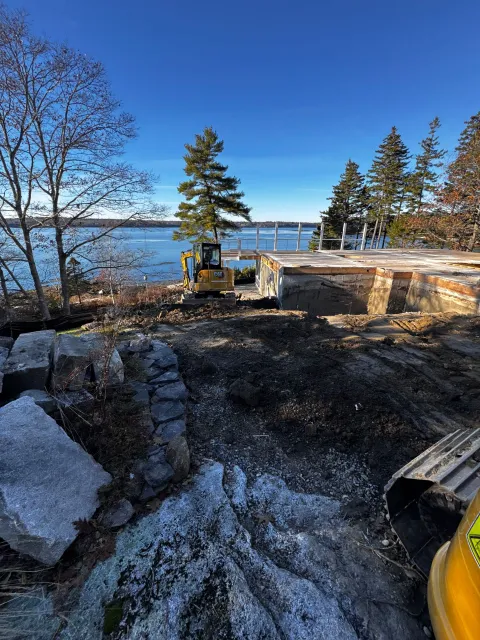 Construction site by water with excavator, foundation, and trees. Sunny day.