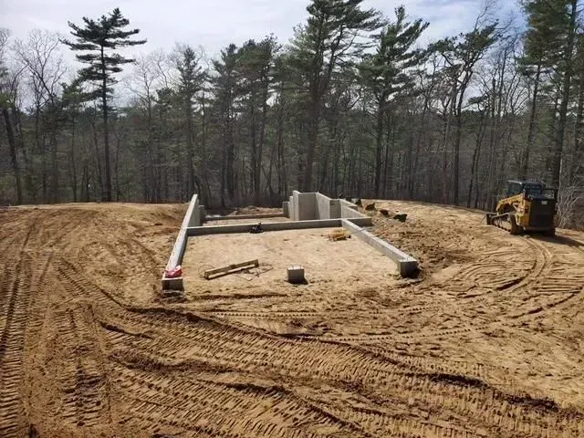 Foundation site with concrete footings, dirt tracks, and a bulldozer in a wooded clearing