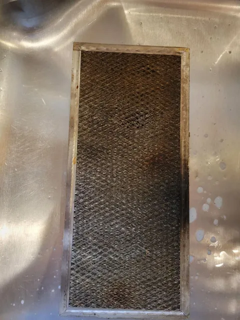 Dirty, rectangular metal range hood filter in a sink, coated in grease.