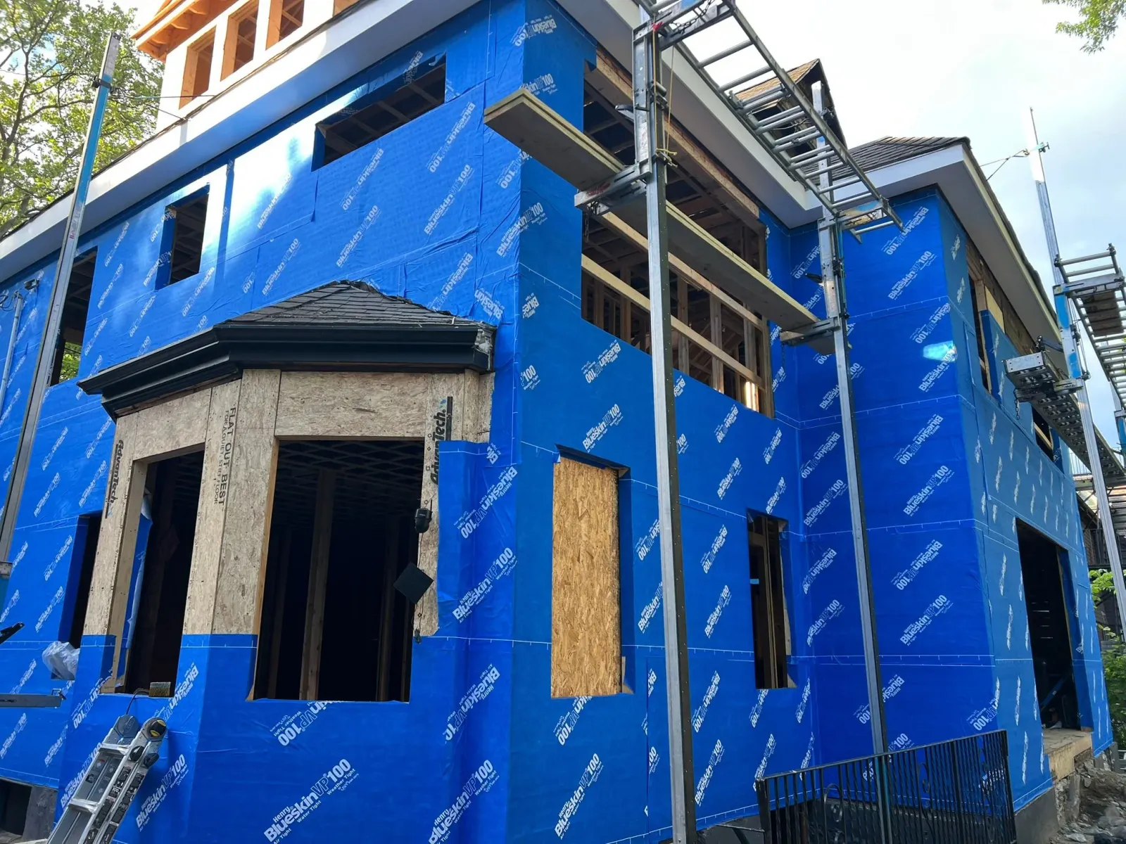 A house under construction with blue sheathing wrap, with exposed wooden framing and scaffolding around it.