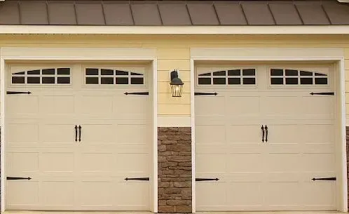 Two beige garage doors with small windows and a wall lantern in between