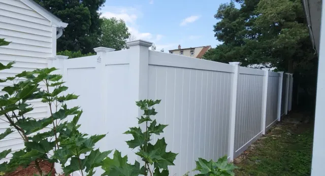 White vinyl privacy fence along a narrow yard beside a house, with trees and cloudy sky.