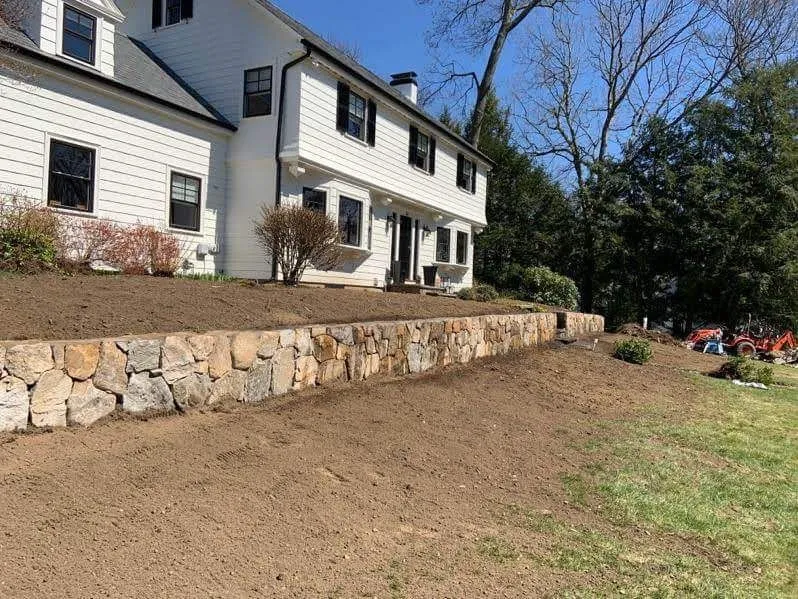 White house with stone retaining wall and sloped dirt yard under a clear blue sky