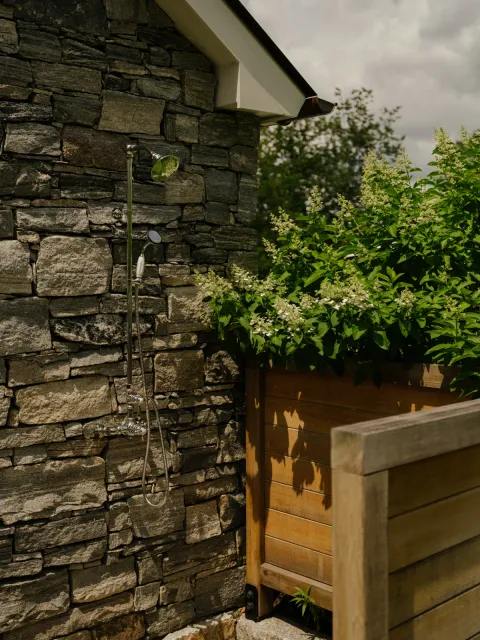 Outdoor shower against a stone wall, with timber screening and flowering shrubs
