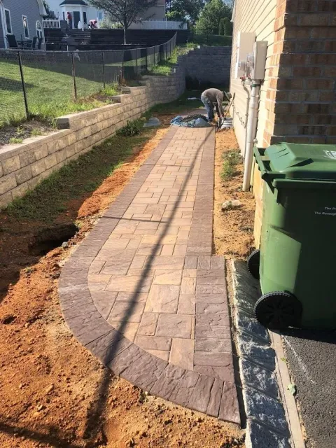 A worker installs a paver walkway along the side of a house, next to a retaining wall and a green trash bin.