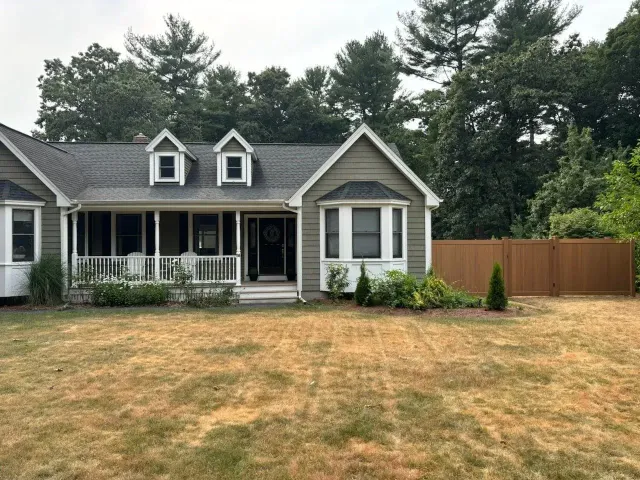 Suburban house with gray siding, front porch, dry lawn, and wooden fence under cloudy skies