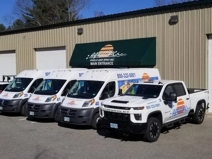 Four white service vans and one white pickup truck parked outside a building with a beige metal exterior and a green awning that reads "Laguna Pools and Spas Inc. Main Entrance." The vehicles have the company's logo, phone number, and colorful sun and wave graphics.