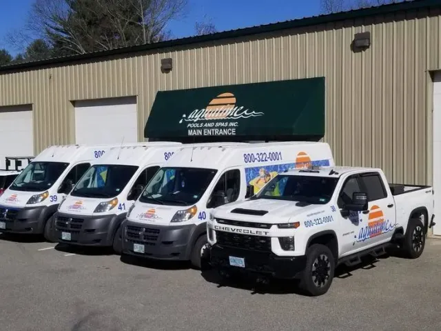 Four white service vans and one white pickup truck parked outside a building with a beige metal exterior and a green awning that reads "Laguna Pools and Spas Inc. Main Entrance." The vehicles have the company's logo, phone number, and colorful sun and wave graphics.