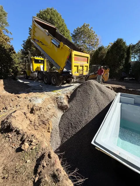 Yellow dump truck unloading gravel into a trench next to a pool shell. A person stands nearby.