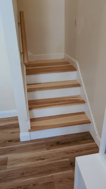 Wooden stairs with white risers in a corner, with a wooden handrail on the left side. The surrounding walls are painted a light neutral color, and a section of the wooden floor is visible at the bottom of the stairs.
