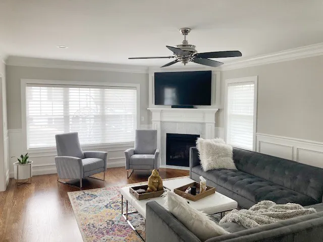 Living room with a stone-look fireplace and TV, windows fitted with white faux-wood blinds, dark gray sectional sofa and armchairs