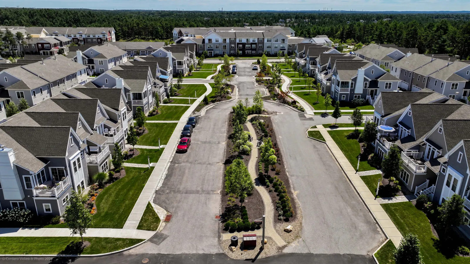 Aerial view of a modern suburban neighborhood with neatly aligned houses, landscaped gardens, and tree-lined streets, surrounded by a forested area.