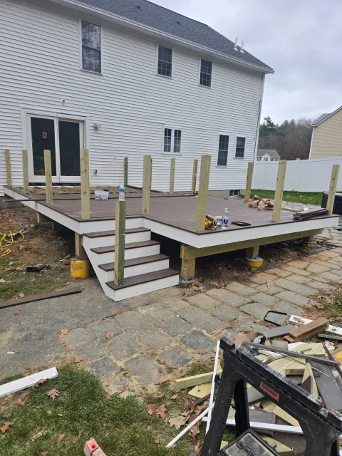 Deck under construction with stairs leading to a backyard. Brown decking and white trim, wooden posts.