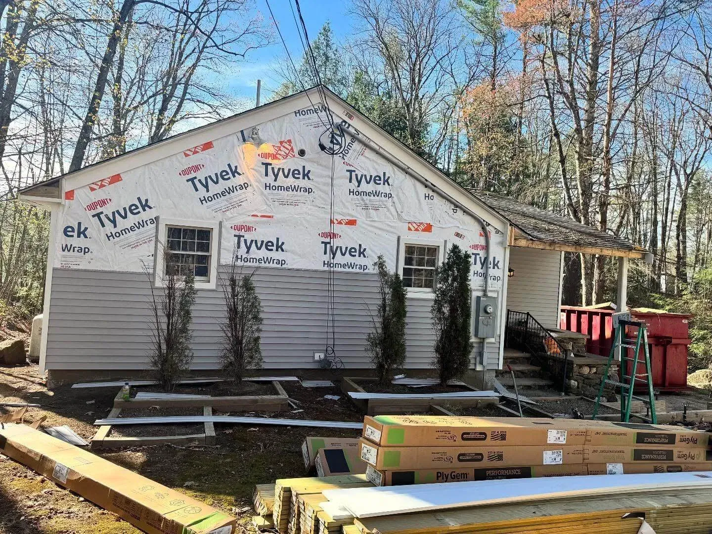 A small house undergoing exterior renovation, with partial Tyvek HomeWrap installed on the upper walls and new gray siding on the lower half. Construction materials and tools are scattered around the yard, and several tall shrubs line the front wall.
