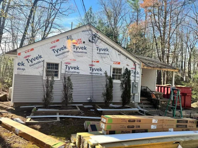 A small house undergoing exterior renovation, with partial Tyvek HomeWrap installed on the upper walls and new gray siding on the lower half. Construction materials and tools are scattered around the yard, and several tall shrubs line the front wall.