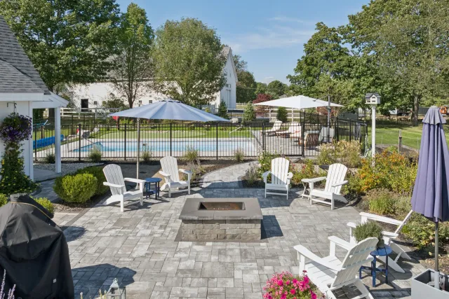Backyard patio with fire pit, white Adirondack chairs, and a pool. Blue umbrellas and green foliage.