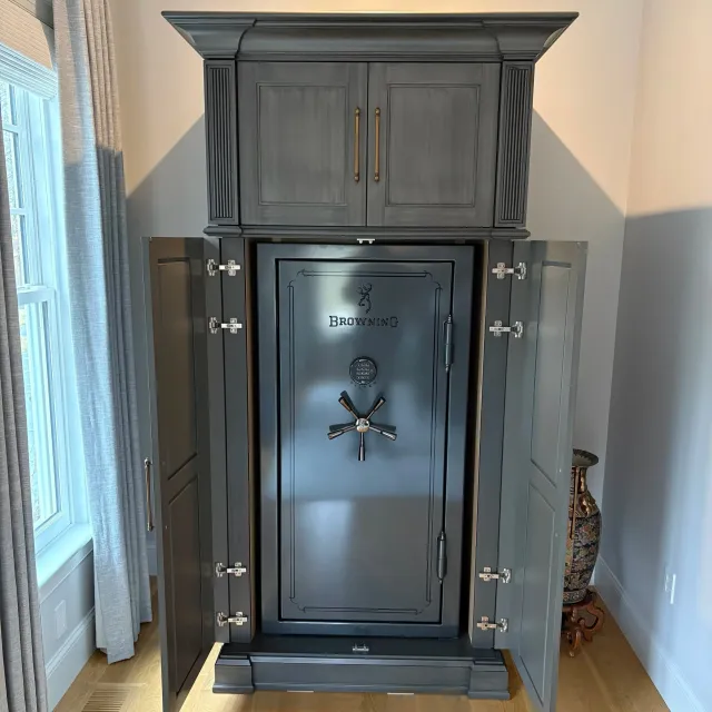 A large, dark gray safe with gold handles and hinges, placed in a room with light-colored walls, hardwood floor, and a window with curtains on the left side.