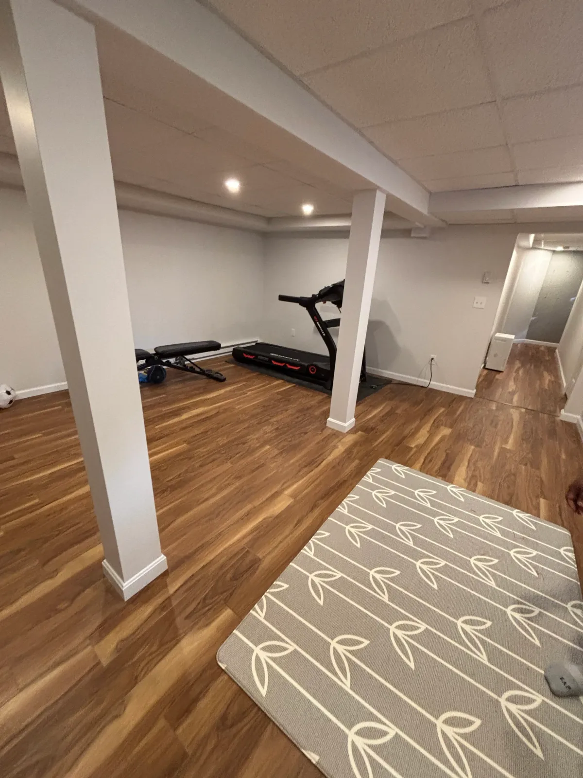 A basement or lower level room with wood flooring, white walls, a treadmill, an exercise bike, a patterned rug, and a doorway.