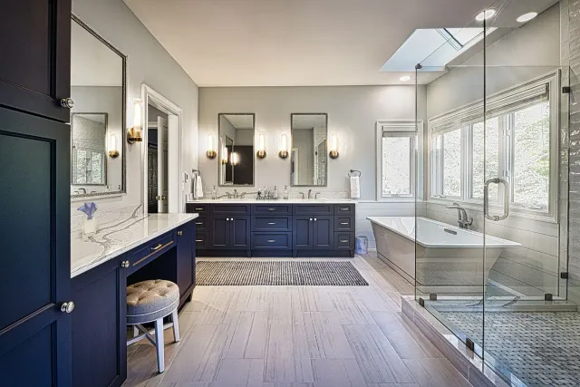 Modern bathroom with navy cabinets, dual sinks, a glass shower, freestanding tub, and large windows letting in natural light.