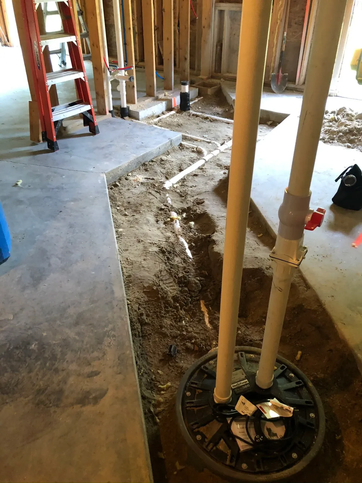 Basement under construction with plumbing pipes, exposed earth, and a ladder. Concrete floor is partially removed for pipe installation.