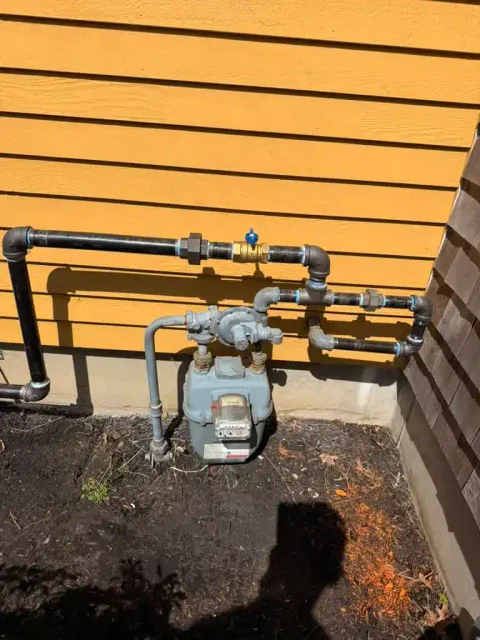 Gas meter with plumbing pipes outside a yellow house.