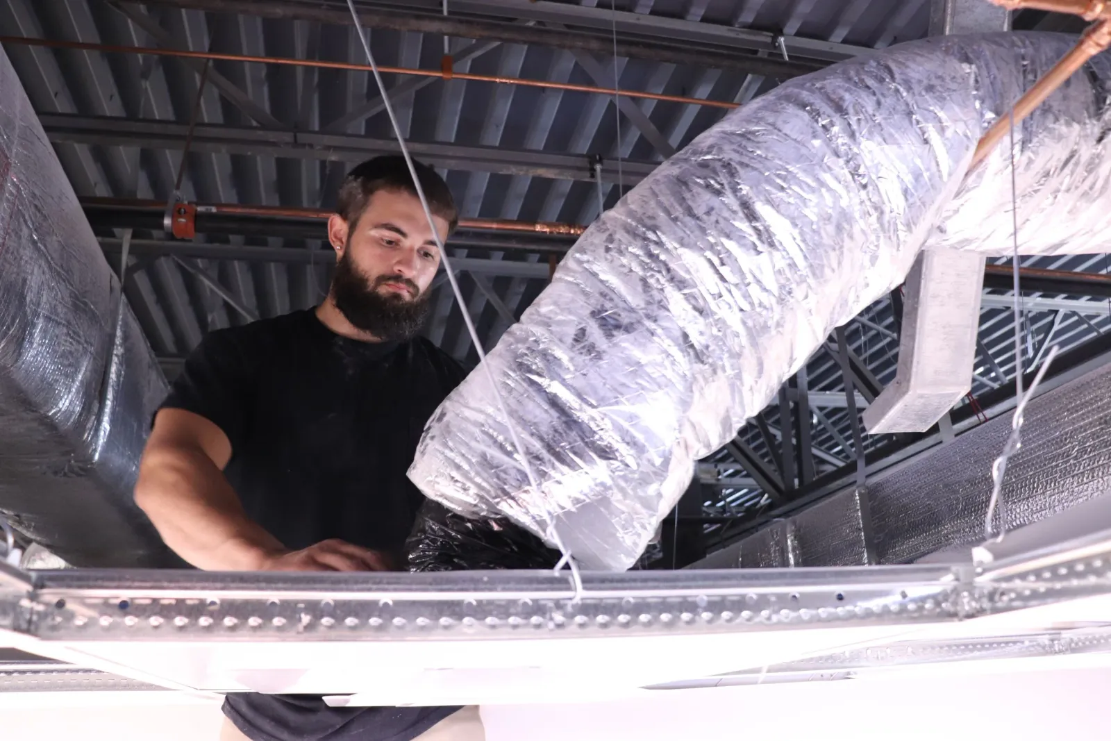 A man with a beard and earrings working in an industrial setting surrounded by large insulated pipes and metal framework.