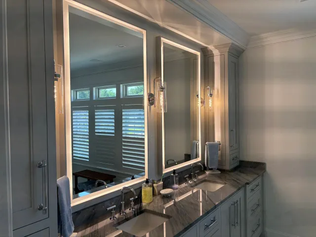 A modern bathroom vanity with two sinks, large illuminated mirrors, and clouds of toiletries. Two wall-mounted lights illuminate the area, and a window with white shutters is reflected in the mirrors.