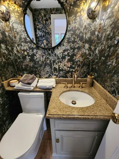 Small bathroom with floral wallpaper, white toilet, granite sink vanity, and oval mirror.