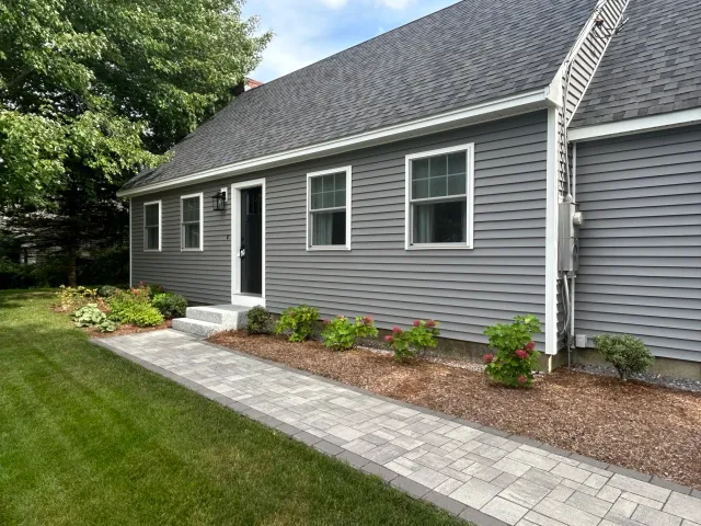 Gray house with white trim, black front door, and shingled roof. Brick walkway leading to entrance, with landscaped garden and green lawn. Tree on the left.