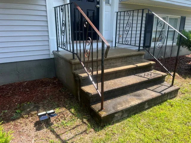 Front steps of a house with black railings and a concrete landing, beside a small lawn light.