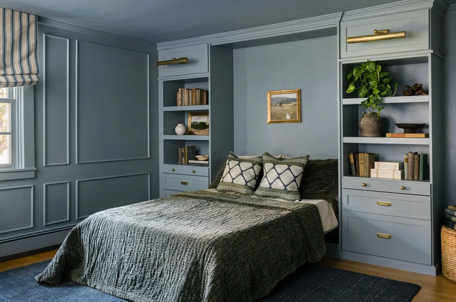 Cozy murphy bed bedroom and office space with built-in bookshelves and green accents, featuring a bed with a quilted bedspread and decorative pillows, framed art, and a window with striped Roman shades. Beautiful millwork on walls lend to historical