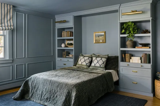 Cozy murphy bed bedroom and office space with built-in bookshelves and green accents, featuring a bed with a quilted bedspread and decorative pillows, framed art, and a window with striped Roman shades. Beautiful millwork on walls lend to historical