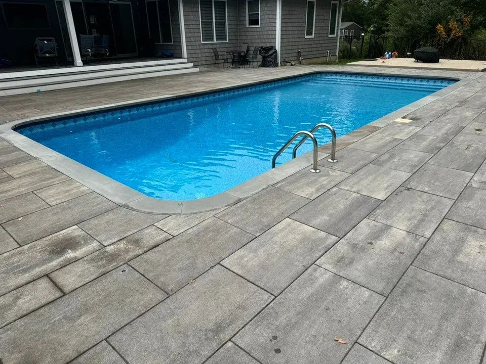 A rectangular swimming pool with blue water and a metal ladder, surrounded by a large grey stone patio beside a house.