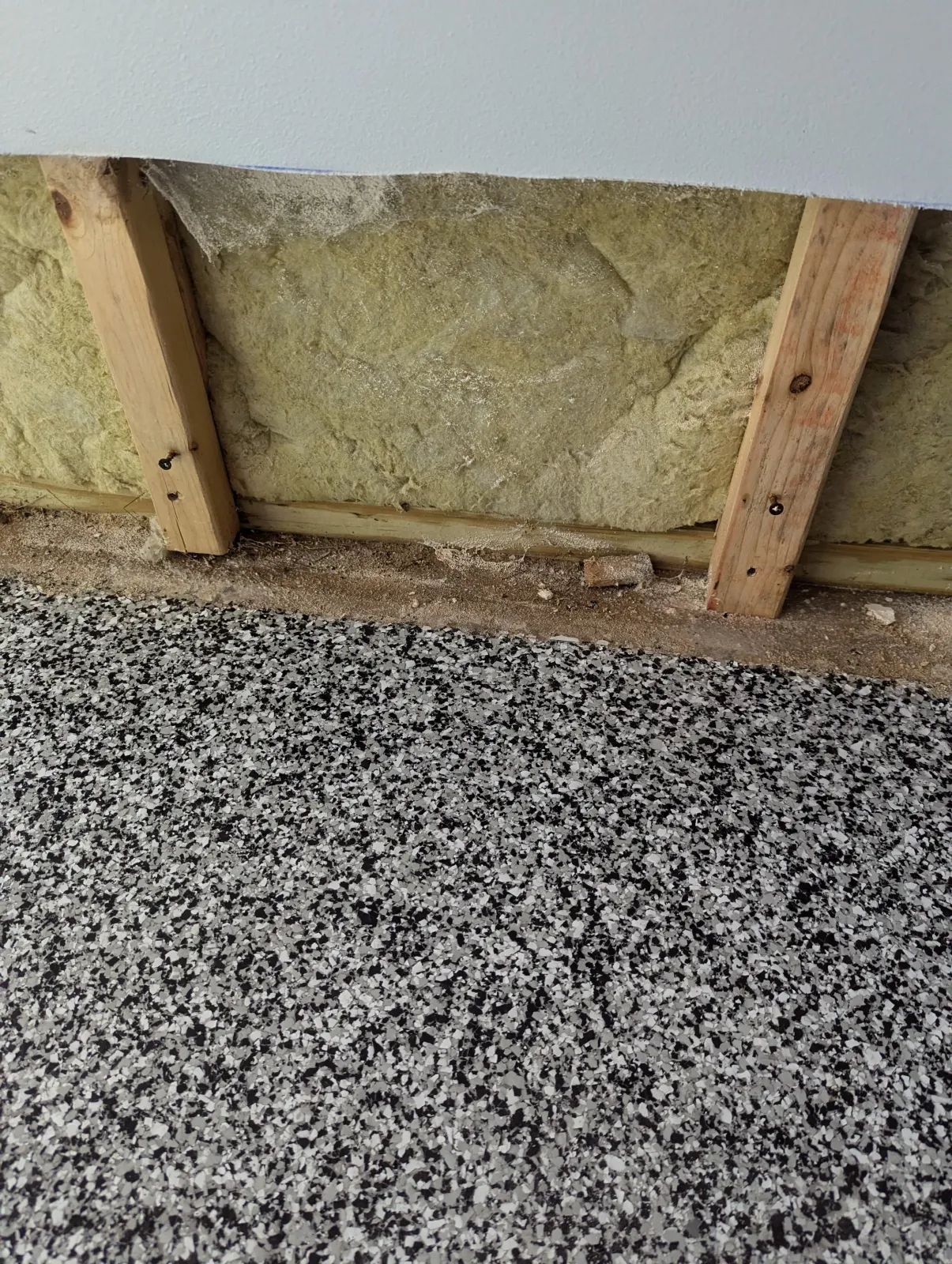 A construction area with insulation material between wooden studs and a speckled black and white floor.