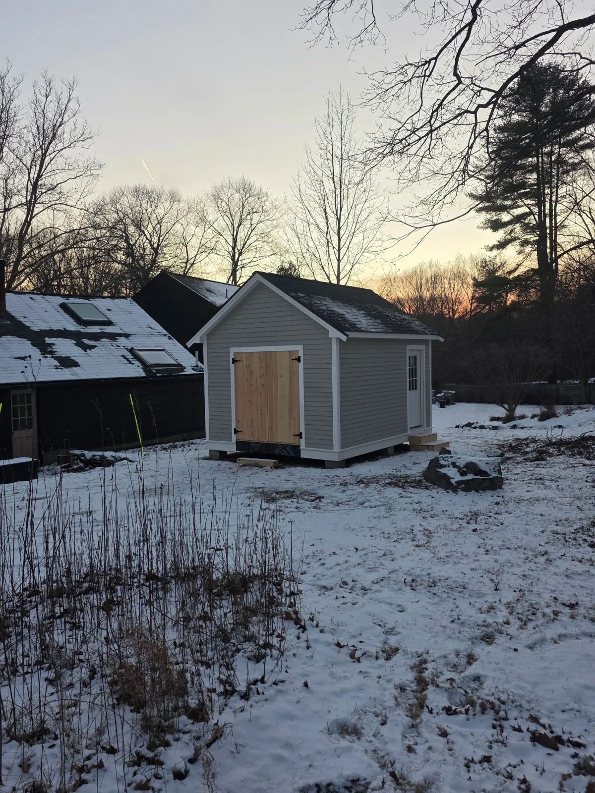 A small gray shed with white trim and a wooden door, situated on a snowy yard, with a large stone nearby, trees in the background, and neighboring shed with a snow-covered roof.