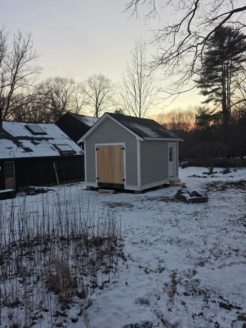 A small gray shed with white trim and a wooden door, situated on a snowy yard, with a large stone nearby, trees in the background, and neighboring shed with a snow-covered roof.