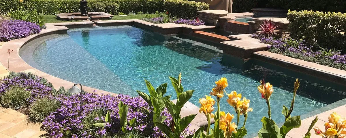 A sunny backyard swimming pool surrounded by purple flowers and yellow canna lilies with stone tiling and a waterfall.