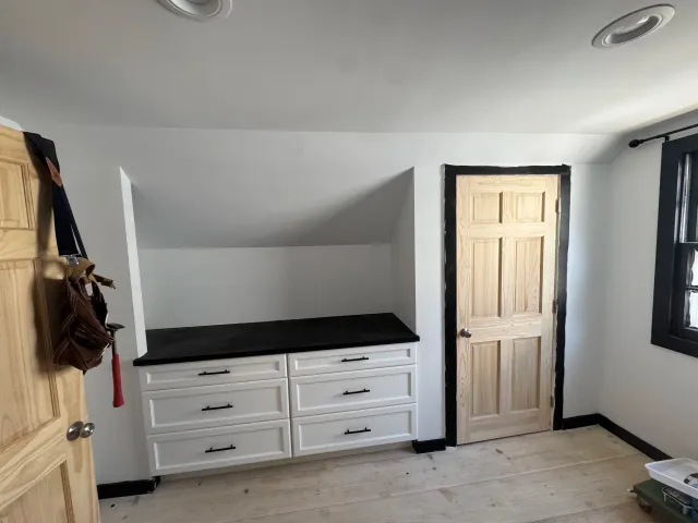 Interior view of a room with white cabinets, black countertop, wooden door, window with black frame, and a sloped ceiling.