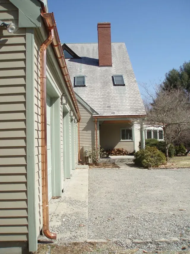 Narrow house side yard with white siding, gravel path, chimneys, and shrubbery under a blue sky