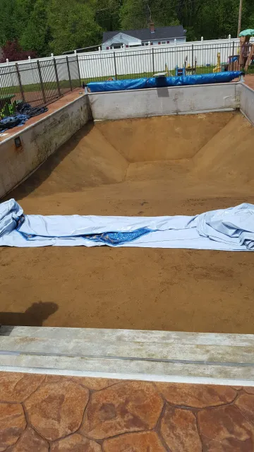 Empty swimming pool with dirt and sand inside, covering part of the pool with a white plastic sheet, located in a backyard with a white fence and green trees in the background.