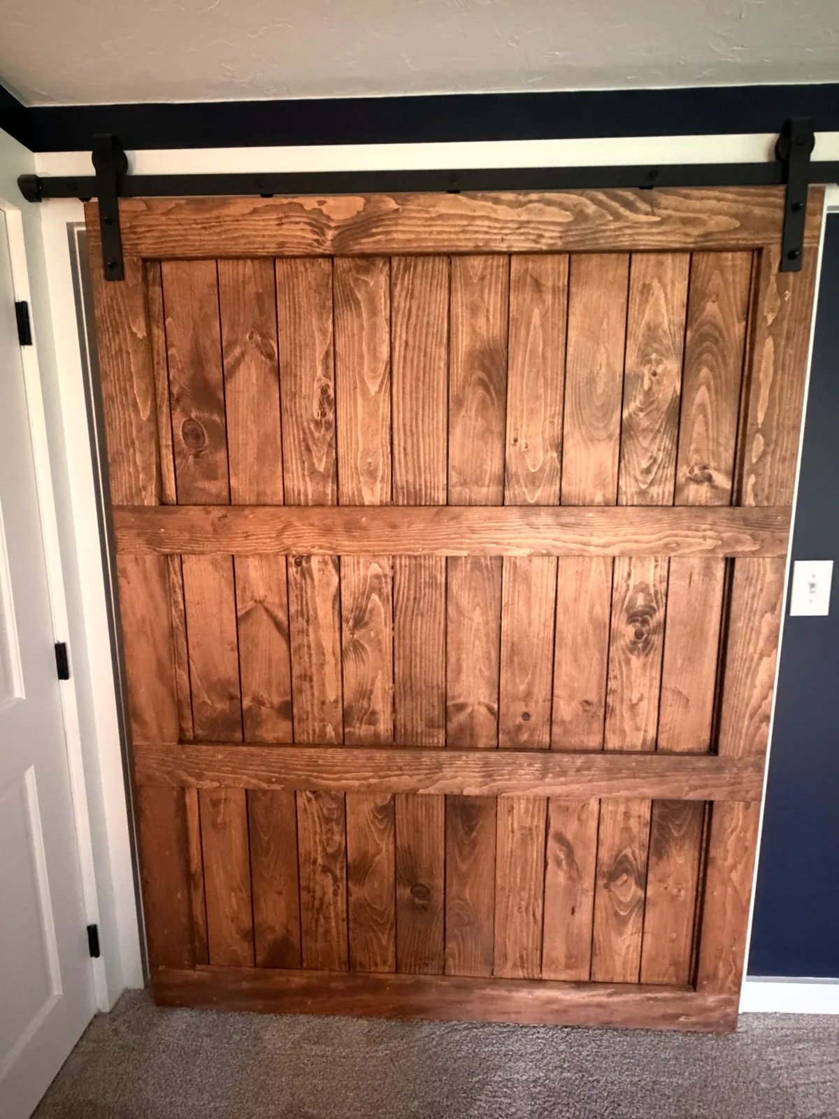 A wooden sliding barn door with black hardware on a wall in a room with a white door and gray walls.