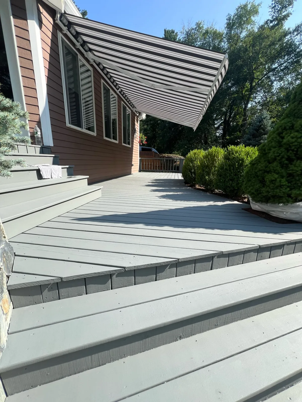 A wooden outdoor deck with stairs, attached to a house, shaded by a striped retractable awning. There are bushes and trees in the background, and a water bottle and towel are on the house steps.