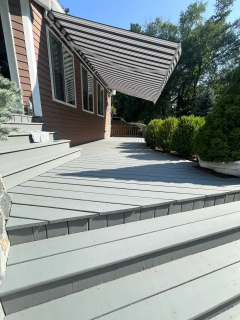 A wooden outdoor deck with stairs, attached to a house, shaded by a striped retractable awning. There are bushes and trees in the background, and a water bottle and towel are on the house steps.
