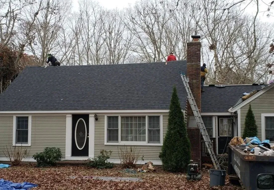 Roofers working on a dark gray shingle roof of a beige house with a black door.