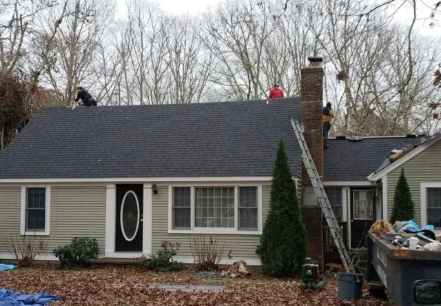 Roofers working on a dark gray shingle roof of a beige house with a black door.