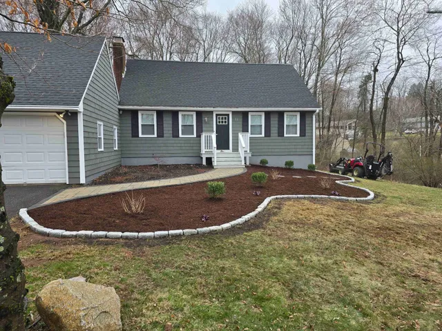 A gray suburban house with a white garage and a fresh, curved mulch garden bed edged with stone pavers.