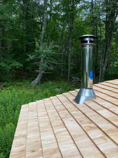 Metal chimney on a wooden roof with a forest background.